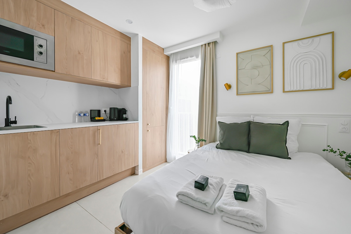 A cozy suite features a double bed with soft linens, complemented by fluffy towels neatly arranged on top. An elegant kitchenette with wooden cabinetry includes a microwave and coffee maker. Artwork adorns the walls, while natural light filters through a nearby window.