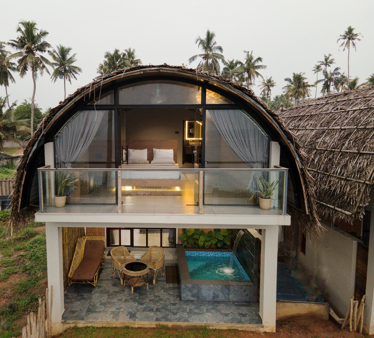 A distinctive architectural structure features a curved thatched roof, with a balcony overlooking a small pool area. Glass doors open to a spacious sleeping area, while palm trees frame the tranquil setting. Outdoor furniture is arranged on the patio, inviting relaxation.