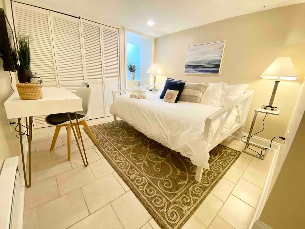 A well-lit sleeping area is presented with a spacious bed dressed in white linens. A light-toned desk is positioned beside the bed, topped with a small plant. A patterned area rug enhances the floor, and soft lighting is provided by lamps on either side.