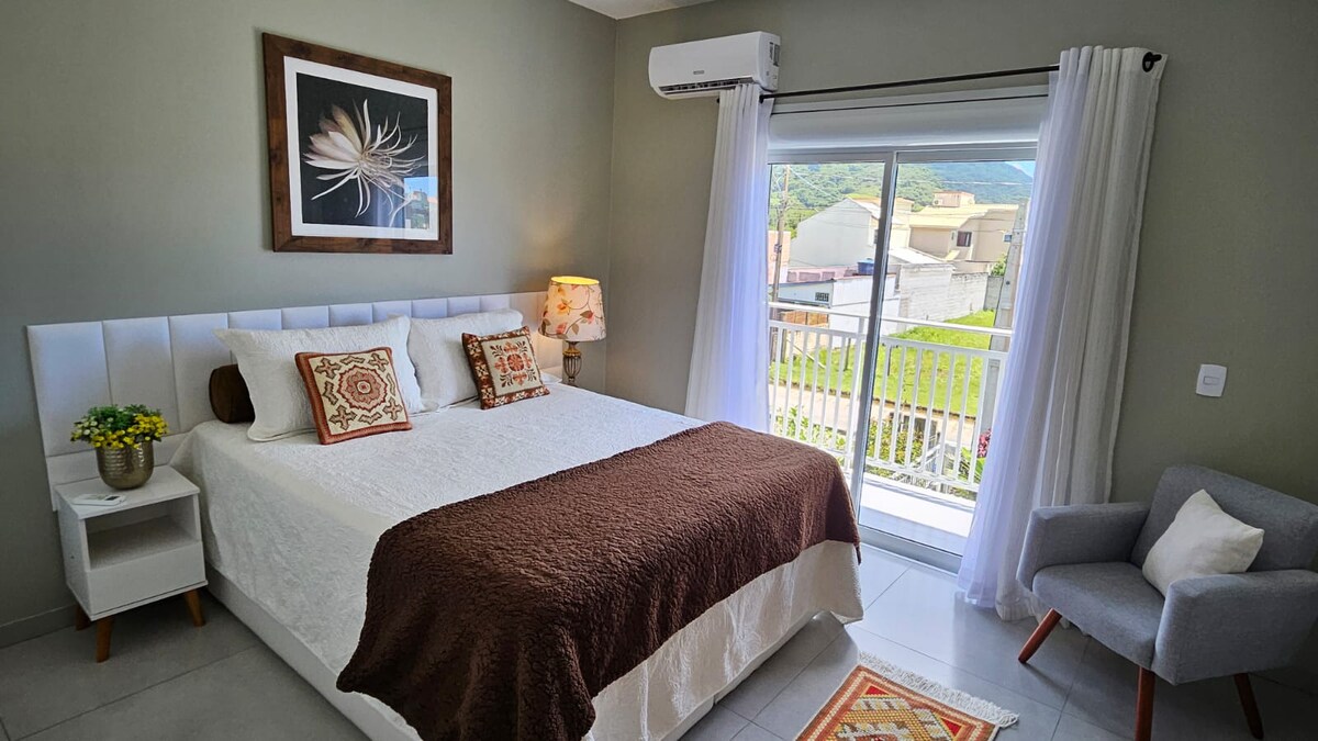 A well-appointed bedroom features a spacious bed with decorative pillows and a chocolate-colored blanket. Natural light floods the room through sliding glass doors that open to a balcony. A small armchair sits near the window, and a floral lamp adds a warm touch.