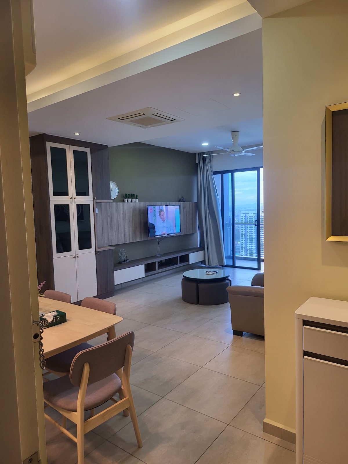 The image captures an open-plan living area showcasing a comfortable seating arrangement with a three-seater sofa and a round coffee table. A wall-mounted TV is viewed from the seating area, and large glass doors allow natural light to fill the space, offering a glimpse of the city views.