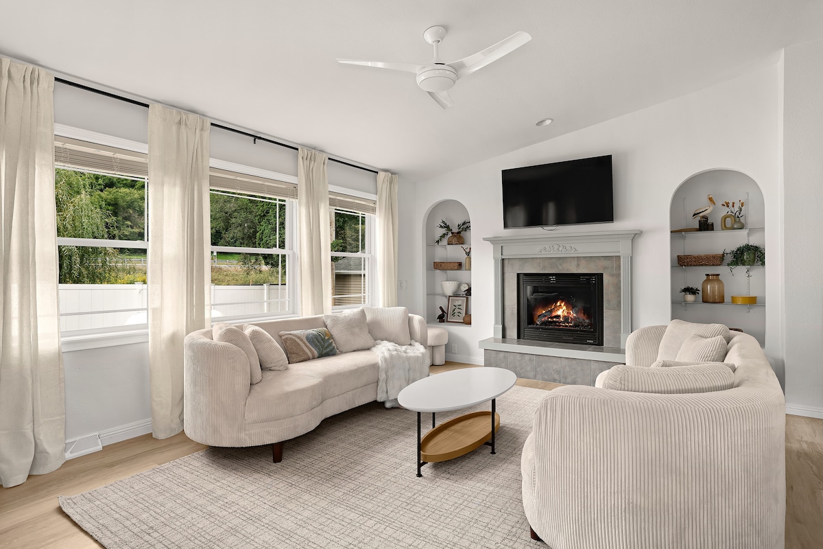 A light-filled living room features soft beige sofas arranged around a circular coffee table. A modern fireplace is set within a gray mantel, with shelves displaying decorative items. Large windows with light drapery provide views of the outdoors, enhancing the room's warm ambiance.