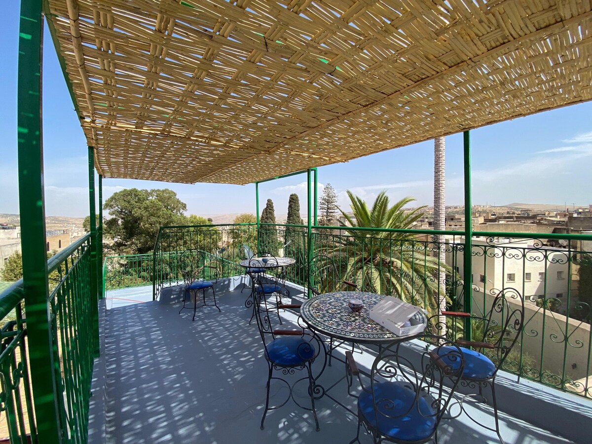 The terrace features a covered area with a woven roof, providing shade. Several tables with intricate tile designs and blue chairs are arranged for seating. Lush palm trees and greenery can be seen in the surrounding views, enhancing the outdoor setting.