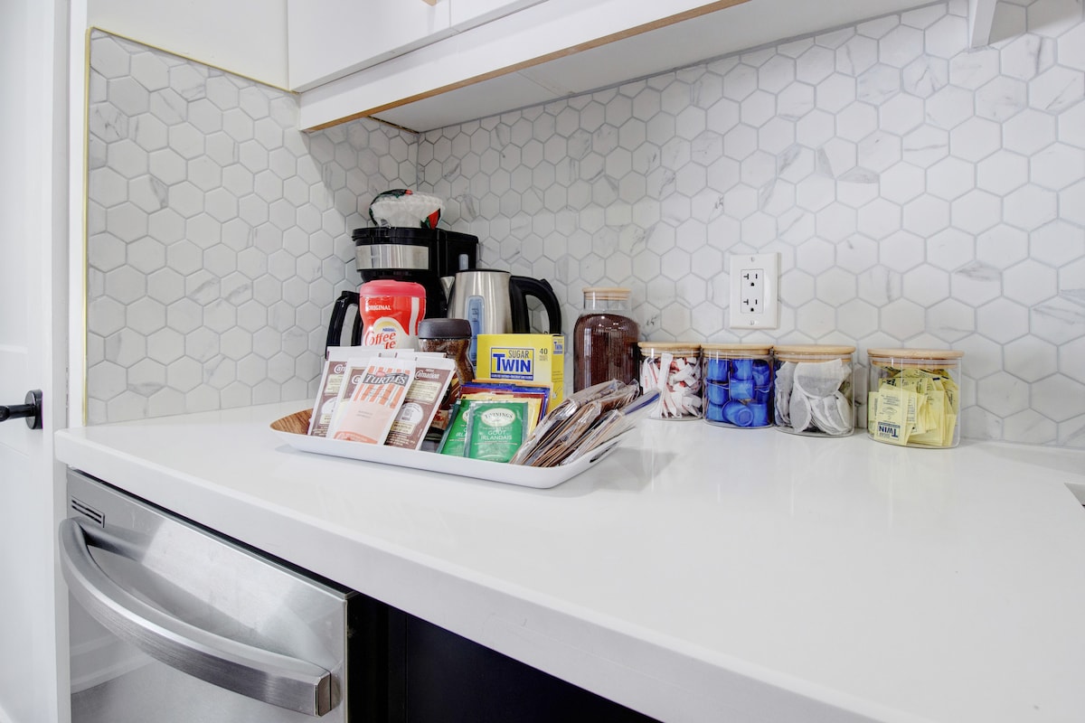 A modern kitchen countertop is featured, showcasing a coffee maker, an assortment of coffee and tea packets, and neatly arranged jars. The surface is adorned with a patterned backsplash of light hexagonal tiles, enhancing the clean and organized appearance of the space.
