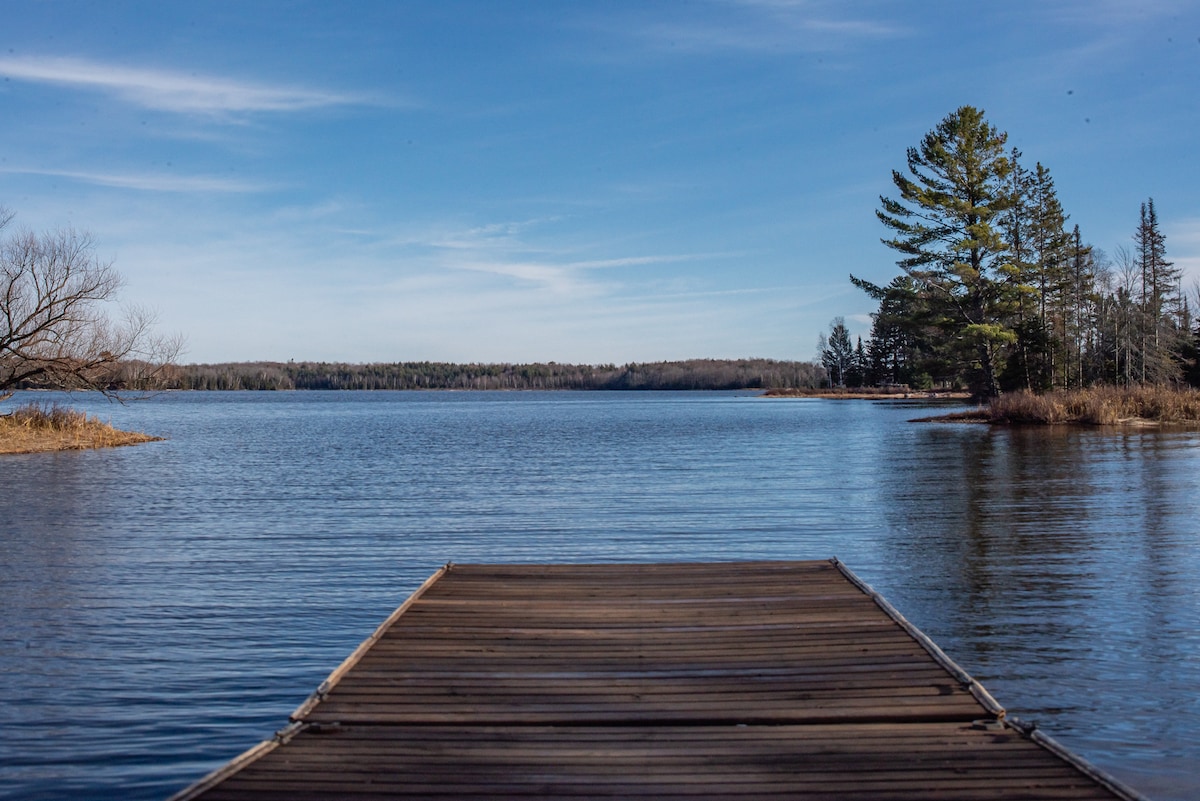 A wooden dock extends over calm waters, leading towards a serene landscape adorned with trees on the opposite shore. The clear blue sky above reflects softly on the water's surface, creating a peaceful scene.