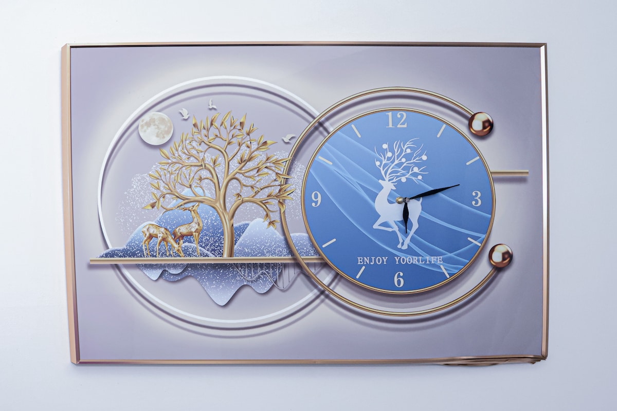 A decorative wall clock features a serene design with a blue face displaying numbers and a silhouette of a couple against a backdrop of golden trees and mountains. The clock is framed with a metallic border, adding an elegant touch to the overall aesthetic.