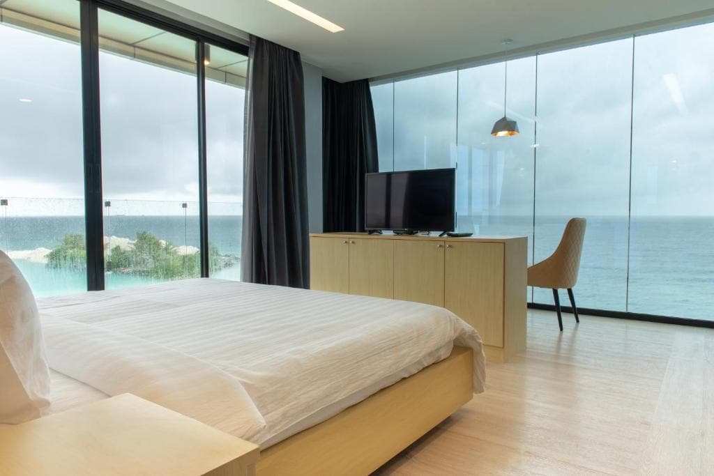 A spacious bedroom features large glass windows offering views of the ocean. A light wood bed is complemented by a simple bedside table. A wall-mounted television is placed on a matching light wood cabinet. A comfortable chair adds to the modern decor.