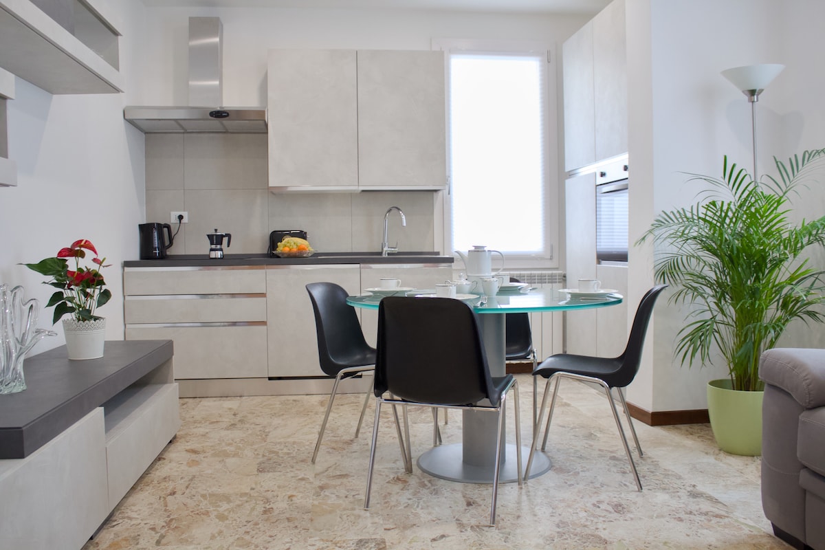 A modern kitchen space is visible, featuring sleek cabinets and a stainless steel oven. A round glass dining table with four black chairs occupies the center. Light enters from a window, illuminating the beige stone floor and a potted plant in the corner.