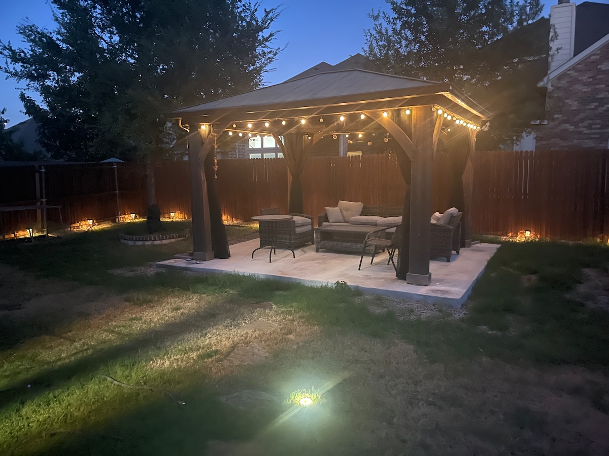 An outdoor pergola is graced with soft café lighting, creating a welcoming space. Comfortable seating is provided beneath the structure, offering a relaxing atmosphere. The surrounding yard features subtle lighting along the perimeter, enhancing the tranquil evening setting.