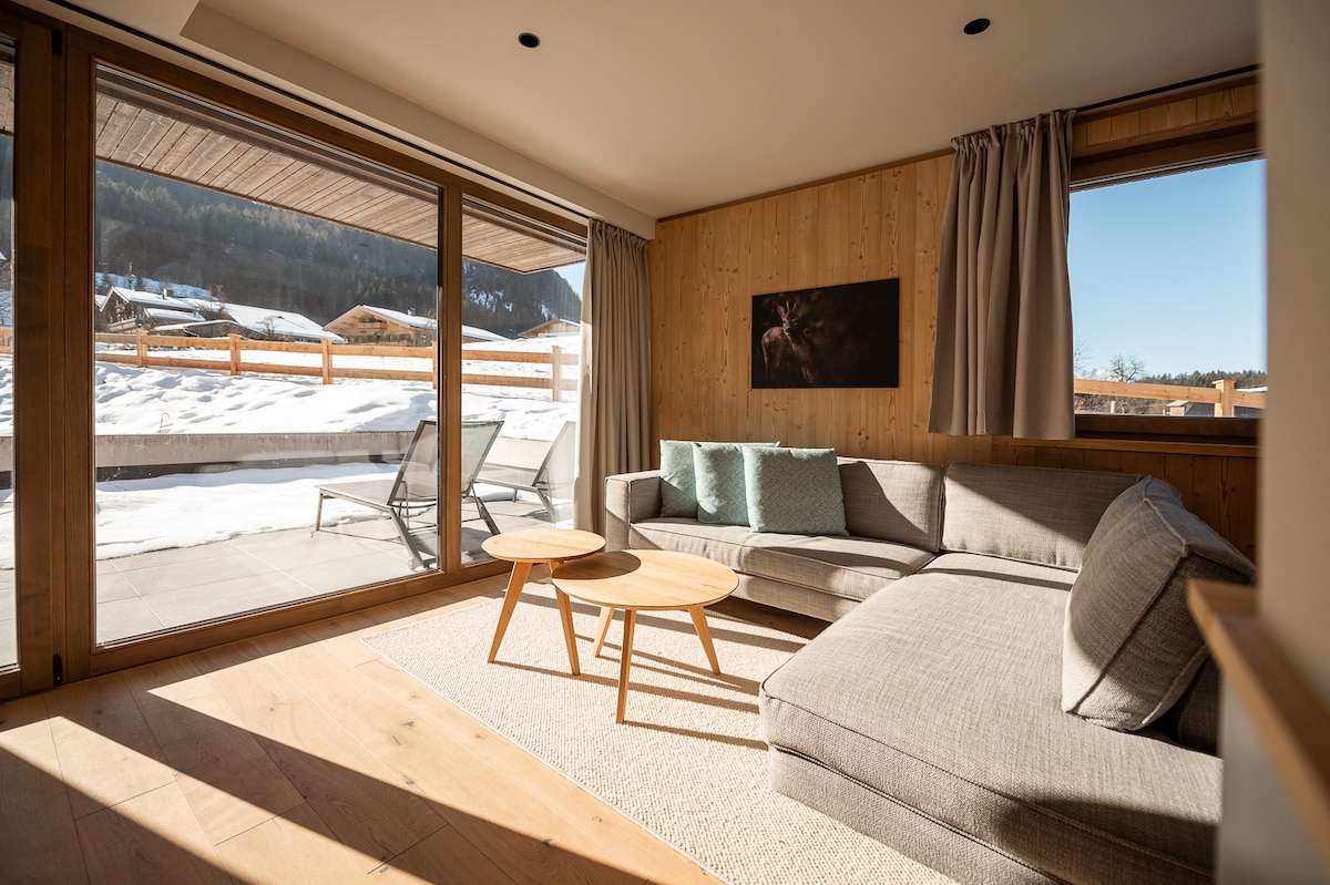 A spacious living area is furnished with a gray sectional sofa and two circular wooden tables. Large glass doors allow natural light to fill the room and provide views of the snowy landscape outside. Neutral tones and wooden accents create a calming atmosphere.