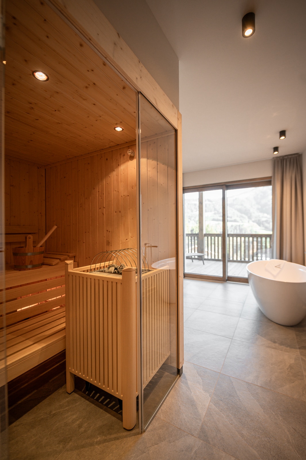 A sauna area features wooden paneling and a transparent glass door, inviting relaxation. Adjacent, a sleek white freestanding bathtub is positioned next to a large window, offering views of the surrounding landscape. Soft lighting enhances the soothing ambiance within the space.