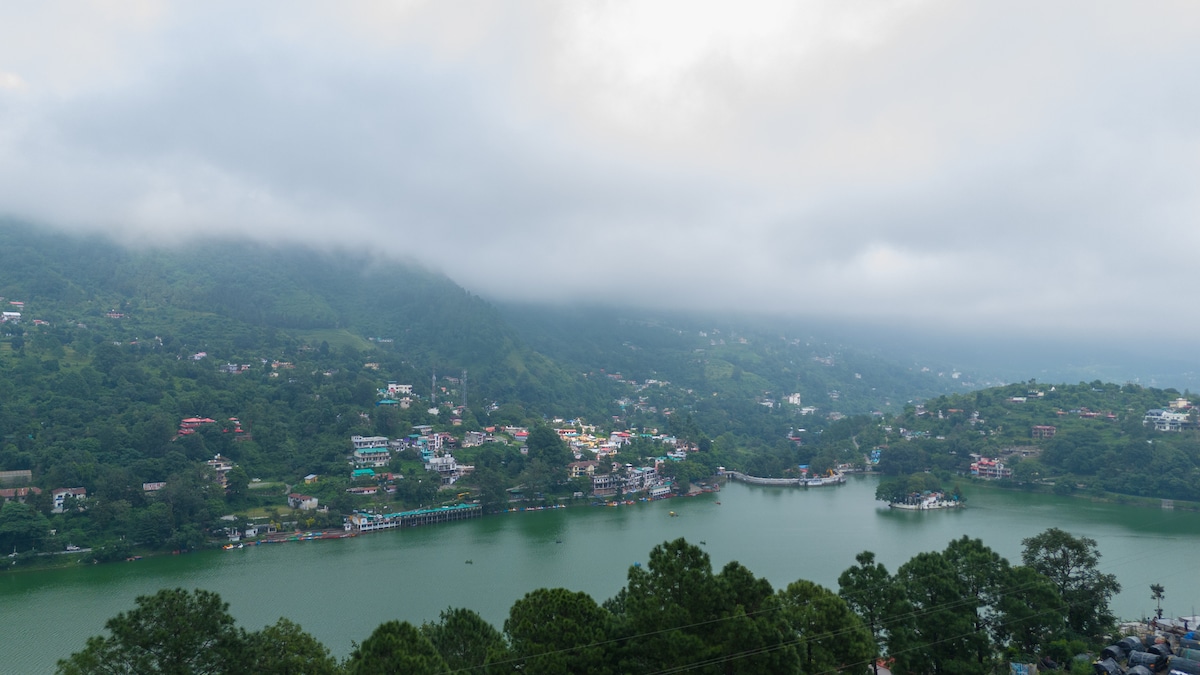 A serene landscape displays a scenic lake surrounded by lush greenery and hills. The water reflects the overcast sky, while small buildings are nestled along the shoreline, creating a tranquil ambience. Mist gently drapes over the mountains, enhancing the natural beauty of the area.
