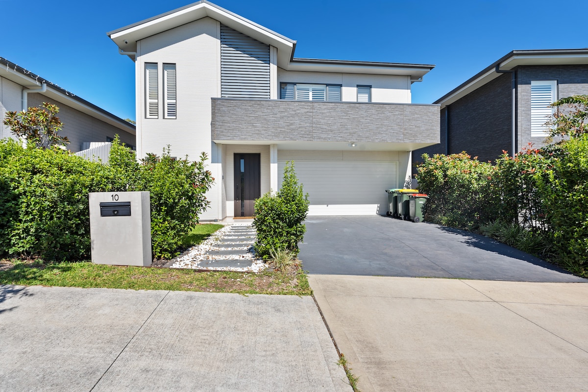 A modern two-story house is presented from a frontal view, showcasing a sleek exterior with a combination of light and dark materials. A tidy driveway leads to a garage, bordered by lush greenery. The front entrance is accentuated by clear lines and large windows, allowing natural light.