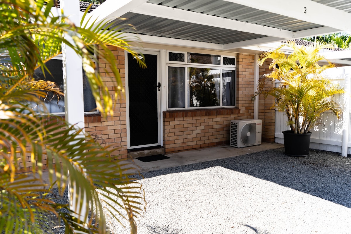 A covered carport provides shelter at the entrance, featuring a large window that allows ample natural light. A small air conditioning unit is positioned next to the entrance, and potted plants add a touch of greenery to the space. The ground is covered with gravel.