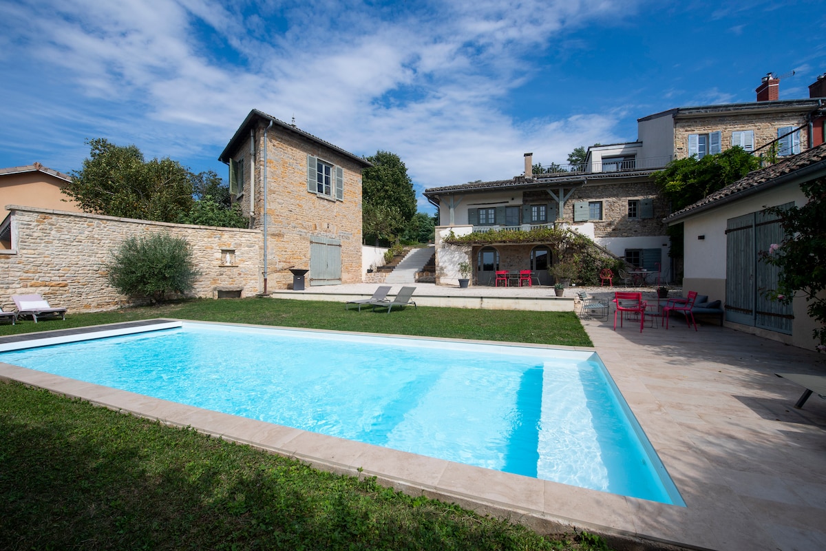 A large heated swimming pool is situated in a green garden area, inviting relaxation. Surrounding the pool are various loungers and outdoor seating. The stone facade of the house and additional structures frame the space, harmonizing with the natural landscape.
