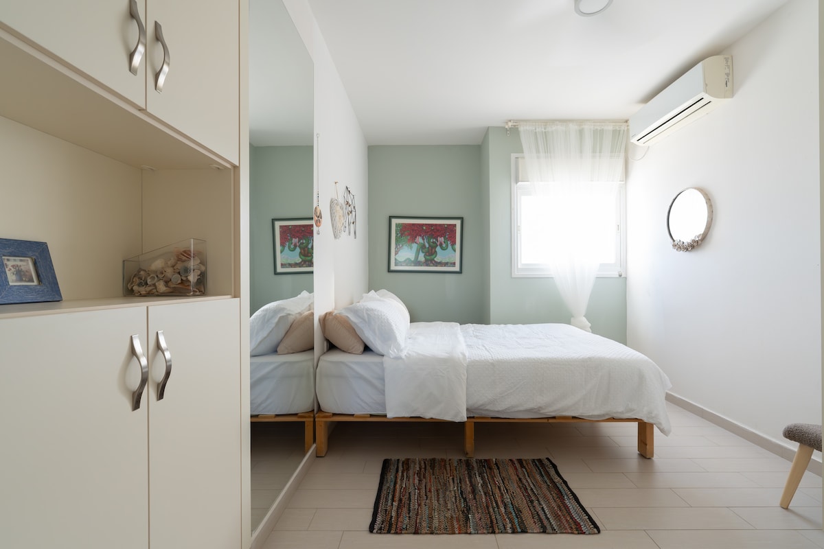 The comfortable bedroom features a spacious bed with crisp white bedding, complemented by soft wall colors. A mirror reflects the decor, and light flows through sheer curtains from a nearby window. A small area rug adds warmth to the wooden floor.