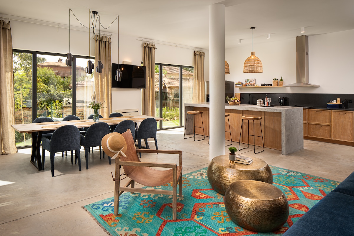 The open-plan living area features a spacious dining table surrounded by dark blue chairs. A cozy lounge area includes light wooden furniture and decorative gold tables. Large windows provide natural light and views of the garden, while neutral tones create a harmonious design.
