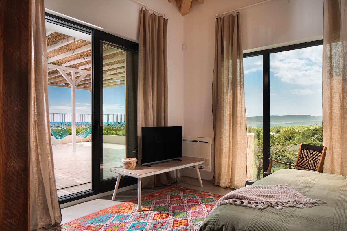 A well-lit sleeping area features a cozy bed with soft bedding, accompanied by a modern television on a white table. Large glass doors open to a spacious terrace, framed by flowing curtains, revealing serene views of the landscape and distant sea.