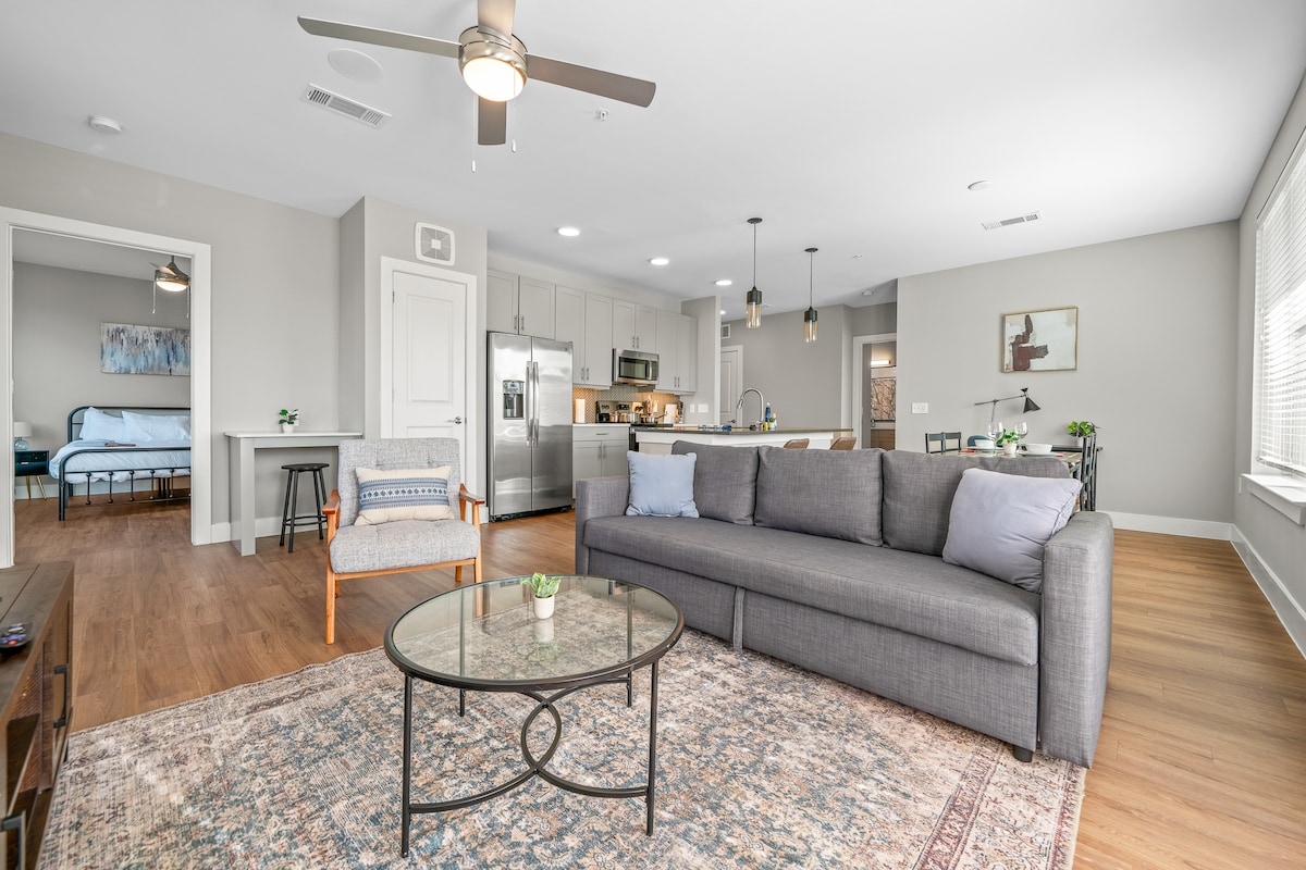 A modern living area showcases a spacious gray sofa, complemented by a circular glass coffee table resting on a patterned area rug. A ceiling fan and large windows enhance the brightness, while an adjacent kitchen area is visible, featuring stainless steel appliances and bar seating.
