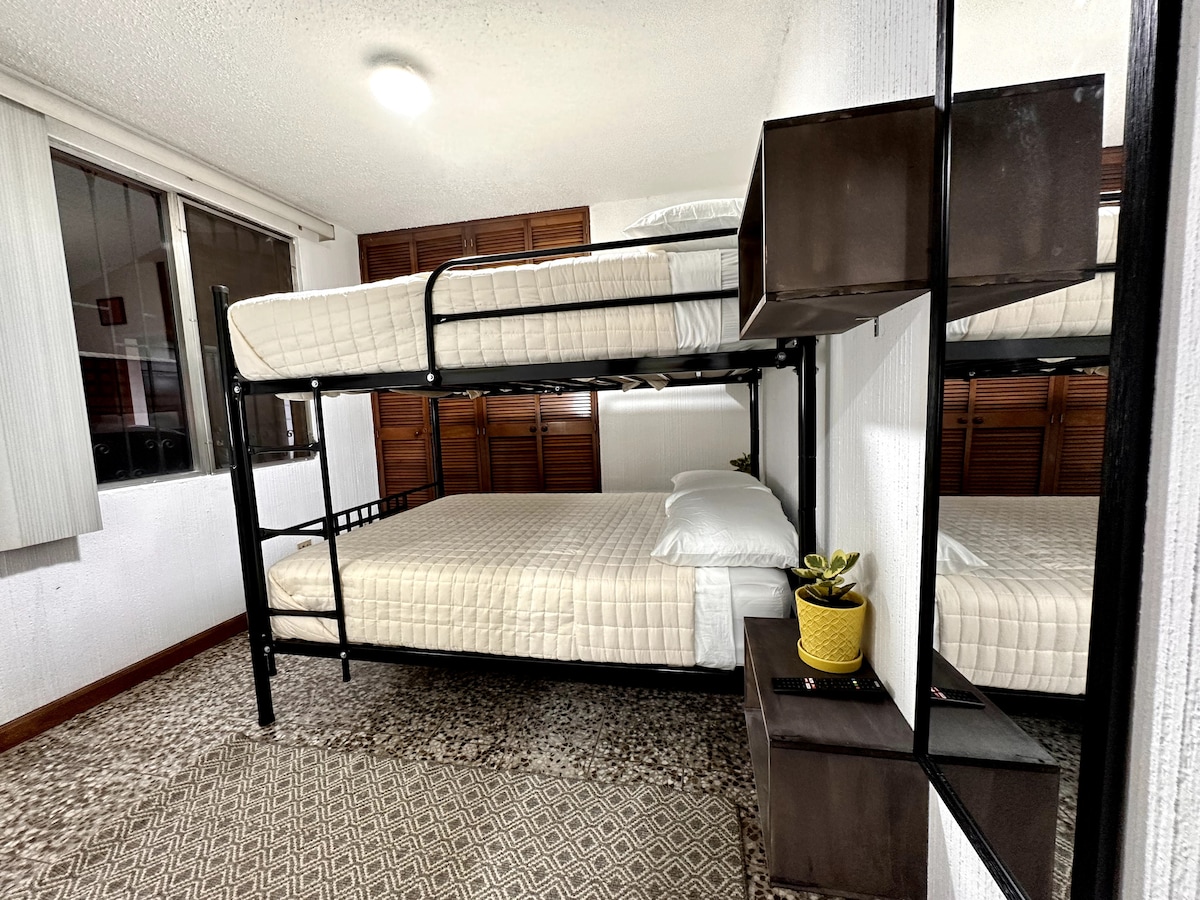 A bedroom features a bunk bed with a spacious bottom sleeping area and a comfortable top sleeping area. Natural light is provided by a nearby window, and a small plant is placed on a bedside shelf. The walls are decorated with neutral tones and wooden furnishings.