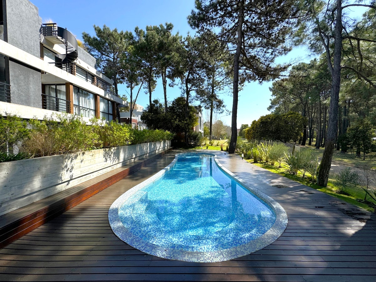 A serene outdoor pool with a gentle curve is surrounded by wooden decking, offering a refreshing blue hue. Tall trees create a natural backdrop, providing shade and enhancing the tranquil setting. Sunlight reflects off the water's surface, inviting relaxation.