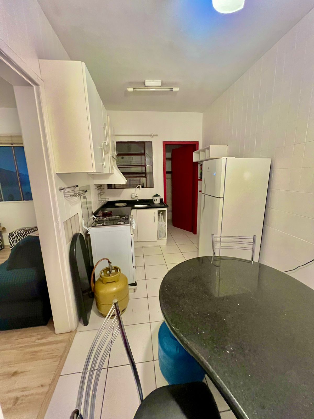 An organized kitchen space features white cabinetry and modern appliances, including a refrigerator and stove. A black countertop is paired with transparent bar stools. Natural light enters through a window, illuminating the room, which also includes a gas cylinder near the floor.