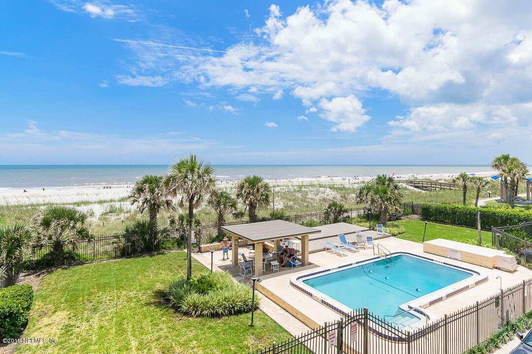 A clear view of the beach is visible from an oceanfront condo, with a pool and sunbathing area in the foreground. Lush greenery surrounds the pool, while the tranquil ocean waves gently meet the sandy shore in the distance under a partly cloudy sky.