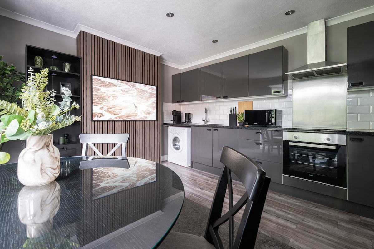 A modern kitchen area features sleek gray cabinetry with an integrated oven and a washing machine. A round glass dining table is accompanied by dark chairs, while a decorative vase with greenery adds a touch of elegance. A wall-mounted TV is visible, enhancing the open layout.