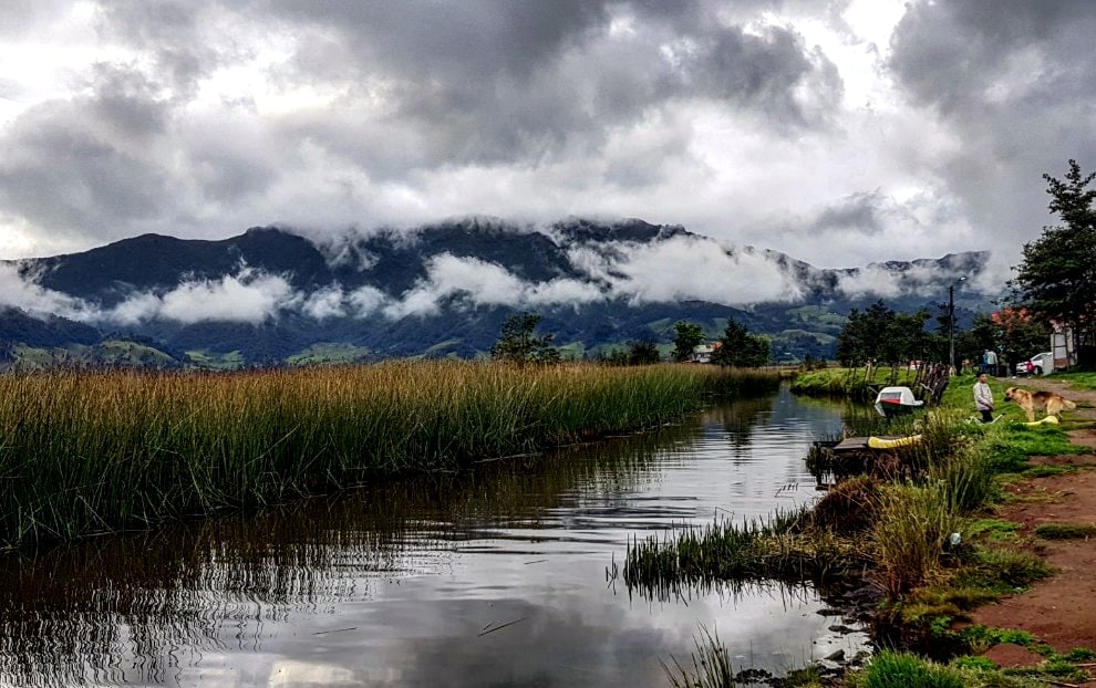 A serene landscape captures a calm waterway reflecting mountainous terrain and cloudy skies. Tall grasses line the banks, and hints of greenery are visible in the distance. The tranquil setting invites peaceful contemplation amidst nature's beauty.