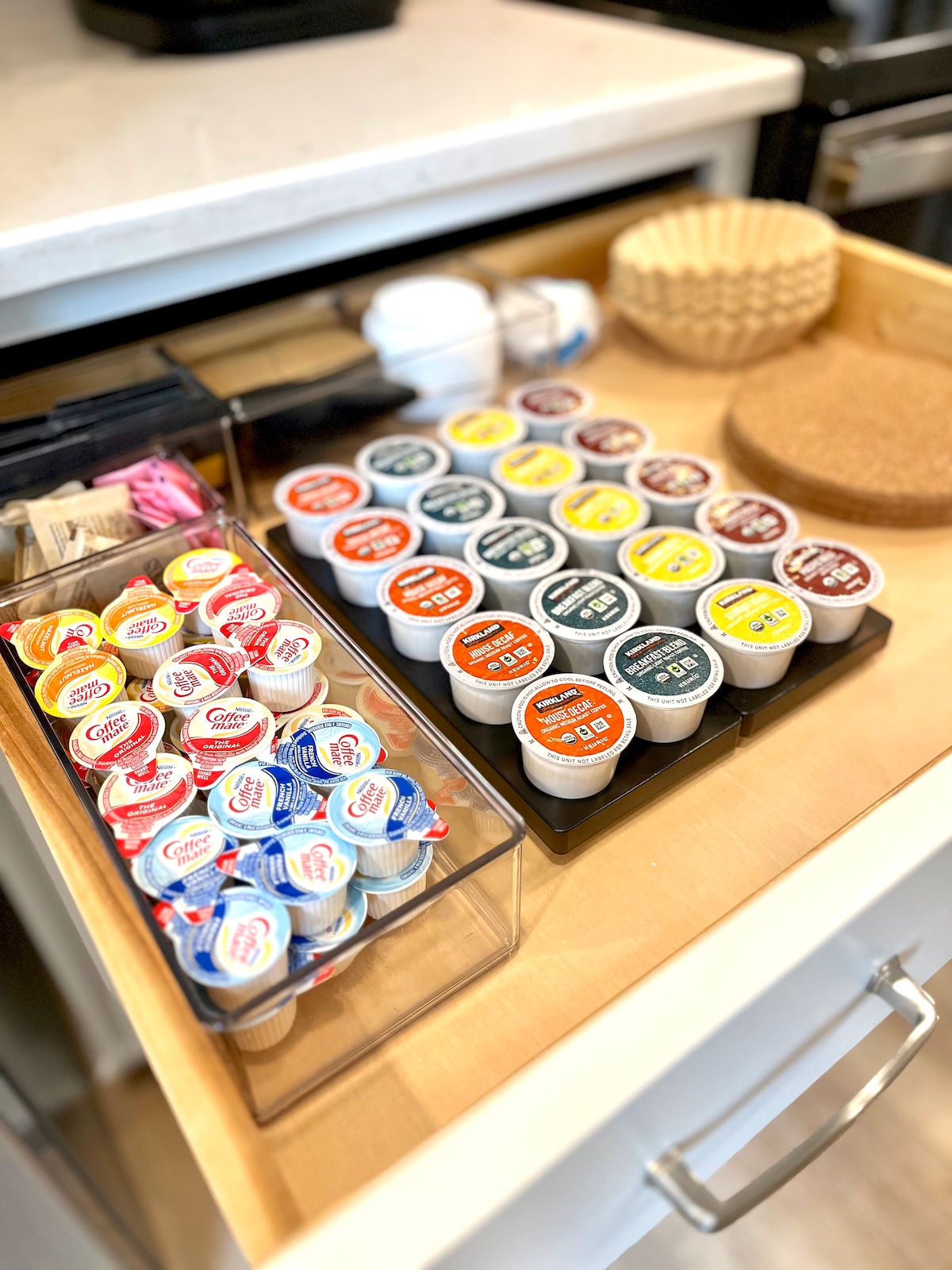 A neatly organized kitchen drawer displays an assortment of coffee and tea options. Various single-serve creamers and flavorings are arranged in clear containers, alongside disposable cups and coasters. The overall setup reflects a thoughtful approach to convenience for guests.