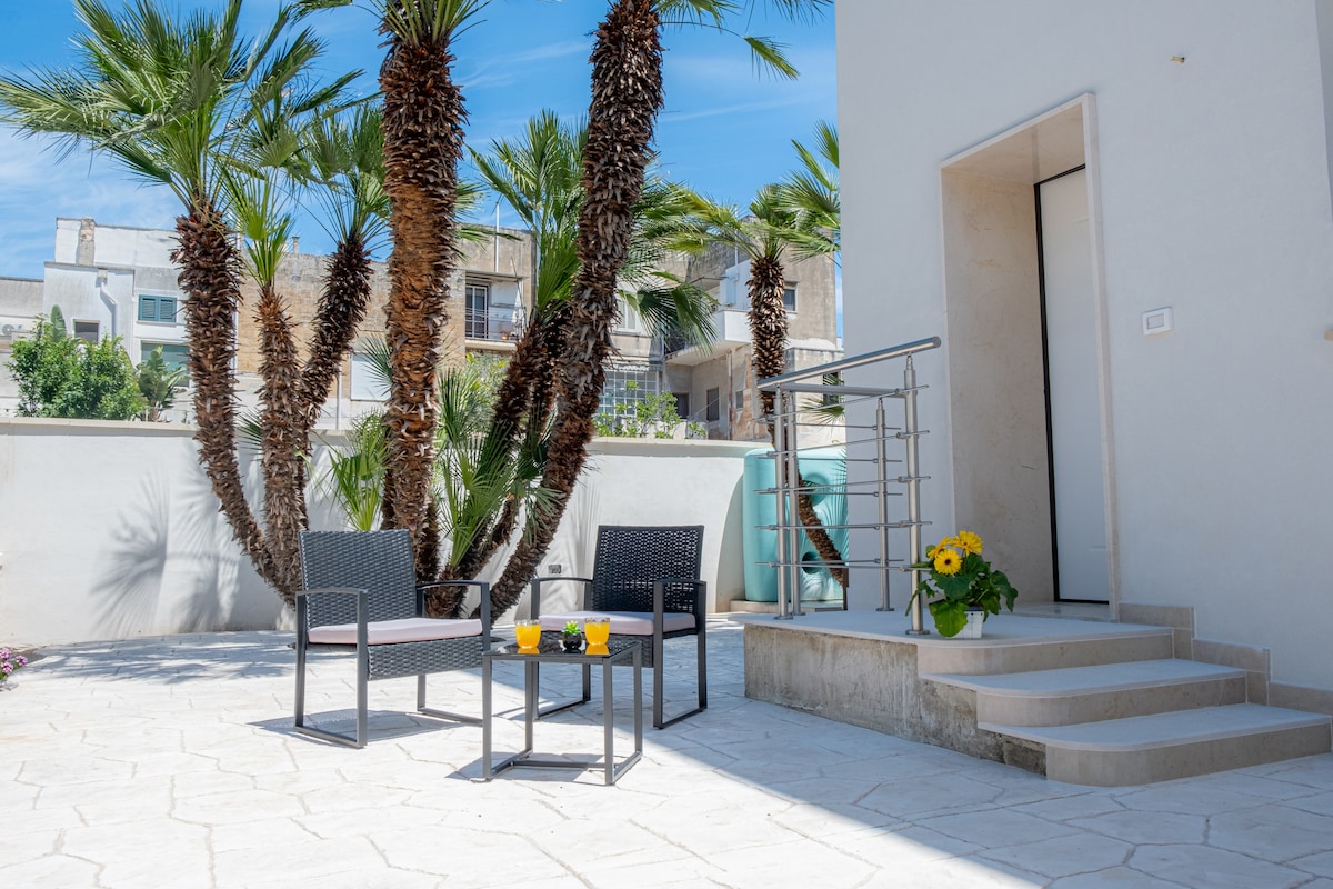 An outdoor area features a light stone patio adjacent to a modern entrance. Two black wicker chairs and a small glass table are arranged nearby, complemented by a pot of yellow flowers. Tall palm trees provide a refreshing backdrop against a clear blue sky.