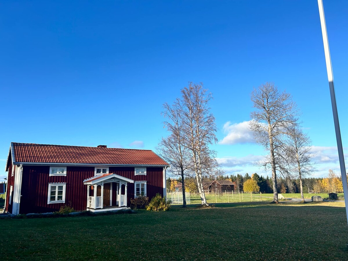 A charming red house is positioned under a clear blue sky, featuring a white entrance and a gabled roof. Surrounding trees are seen alongside a well-maintained lawn, enhancing the rural ambiance of the location.