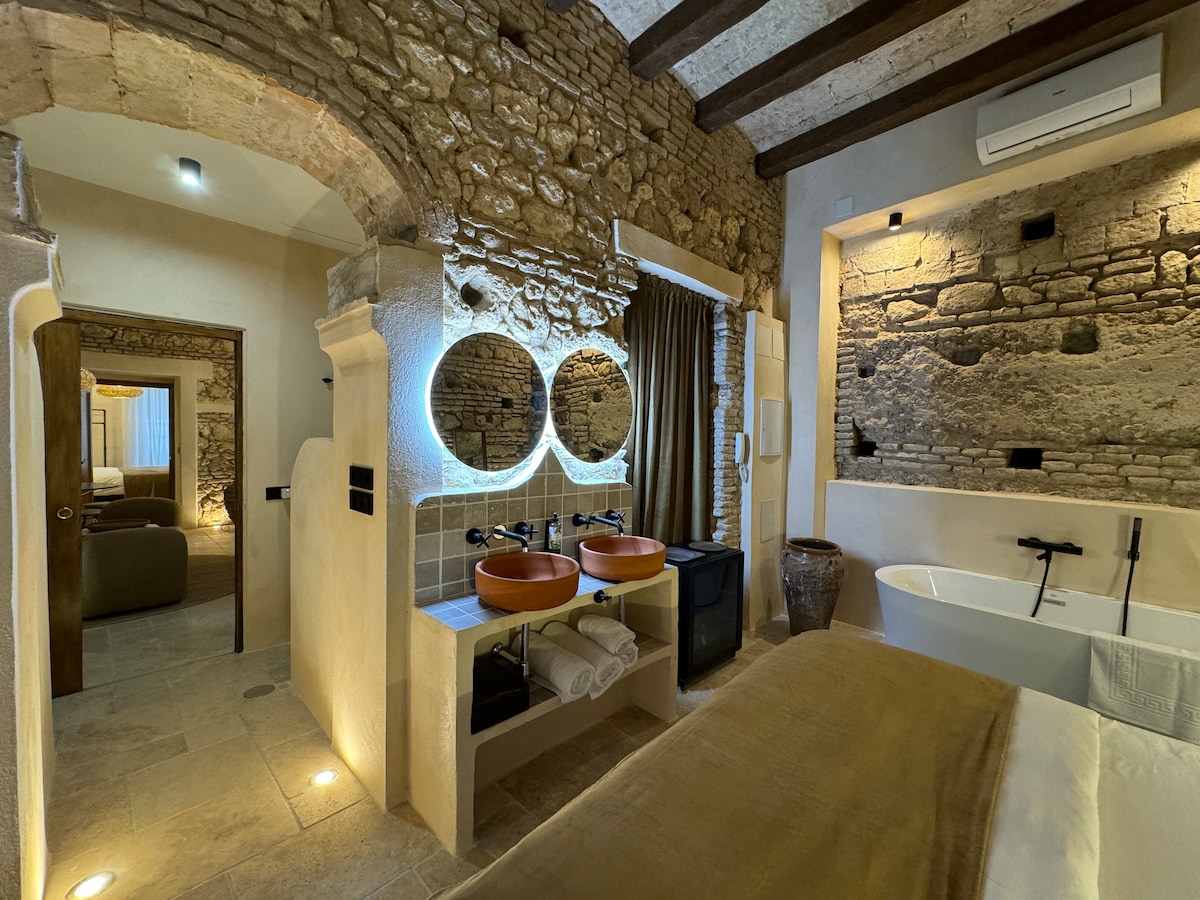 The bathroom features dual circular mirrors above striking terracotta sinks, complemented by natural stone walls. A modern freestanding tub is positioned beside the sinks, with soft lighting enhancing the rustic charm. A cozy bedroom can be glimpsed through an archway in the background.