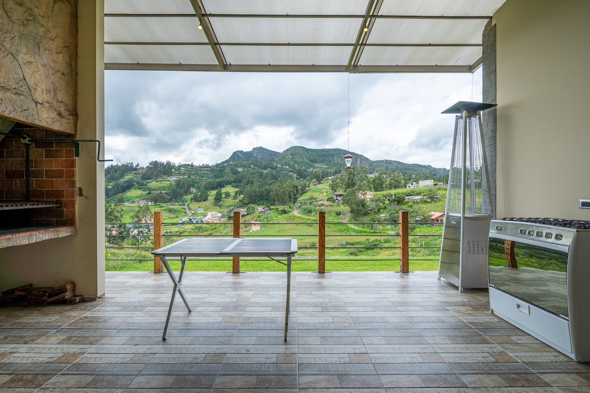 An open kitchen area features large glass panels that provide a clear view of the surrounding mountains. A gas stove and a table are positioned nearby, while a grill is located to the left. The spacious area is designed for social gatherings and outdoor dining.