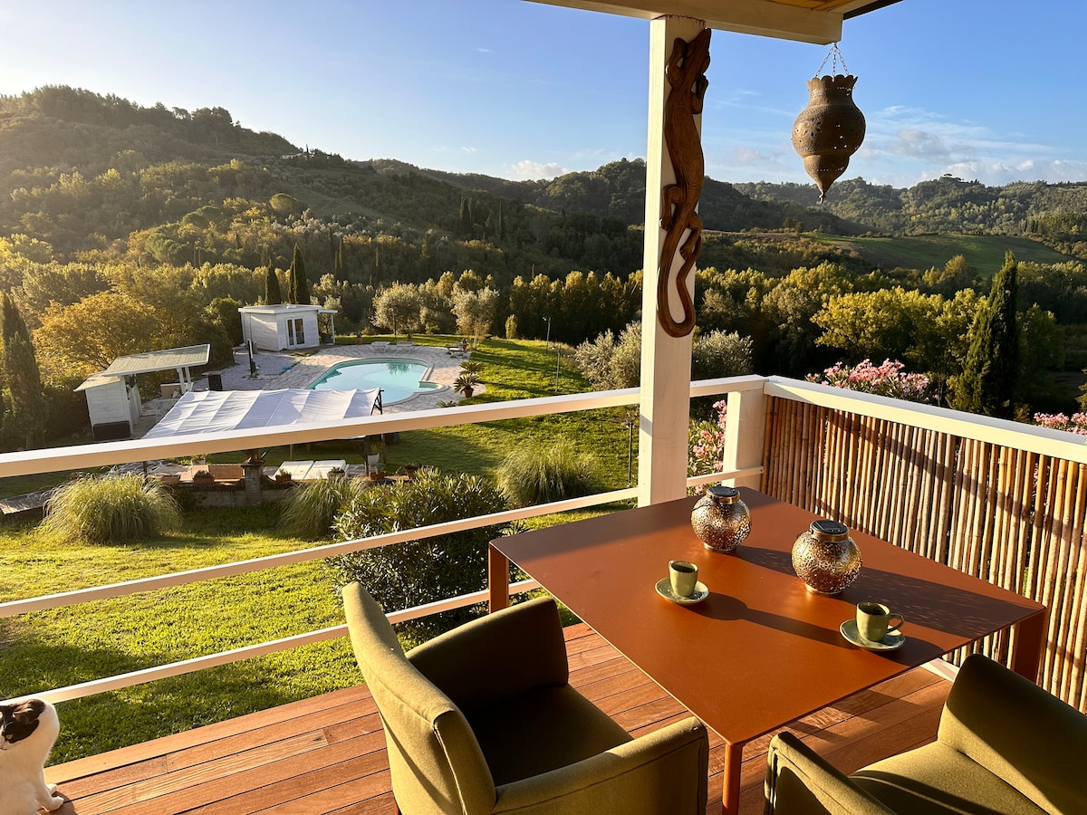 A private terrace features a table with two chairs and decorative objects. The surrounding landscape reveals lush hills and a swimming pool in the distance, complemented by a clear sky and soft sunlight. The vibrant greenery adds a natural touch to the serene setting.