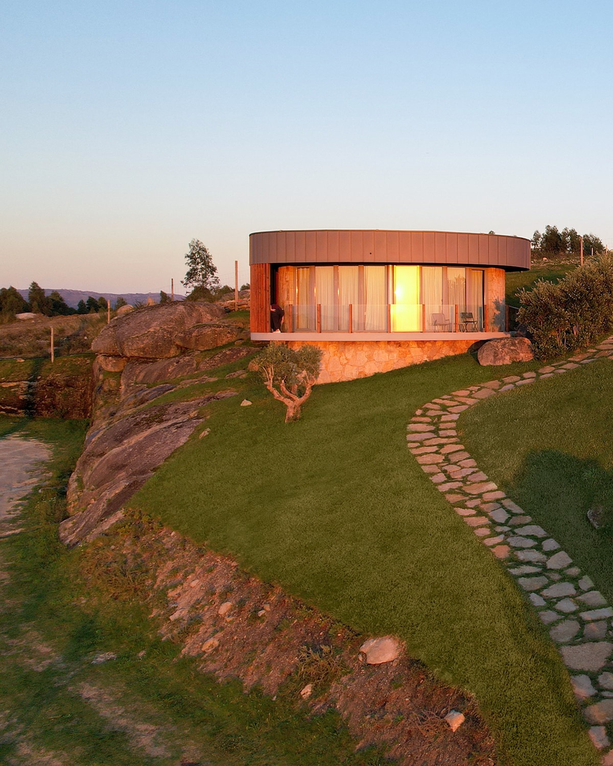 A modern round villa is positioned on a hillside, featuring large glass windows that reflect the warm glow of sunset. A stone pathway winds through the lush green lawn, leading toward the entrance. Surrounding trees and rocky formations add to the natural setting.
