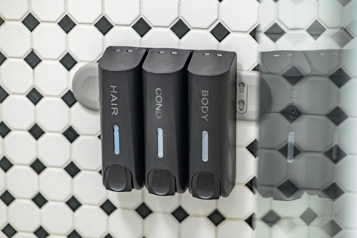 Three dispensers are mounted on a wall in the shower area, labeled for hair, conditioner, and body wash. The dispensers are sleek and black, positioned against a backdrop of classic black and white hexagonal tiles.