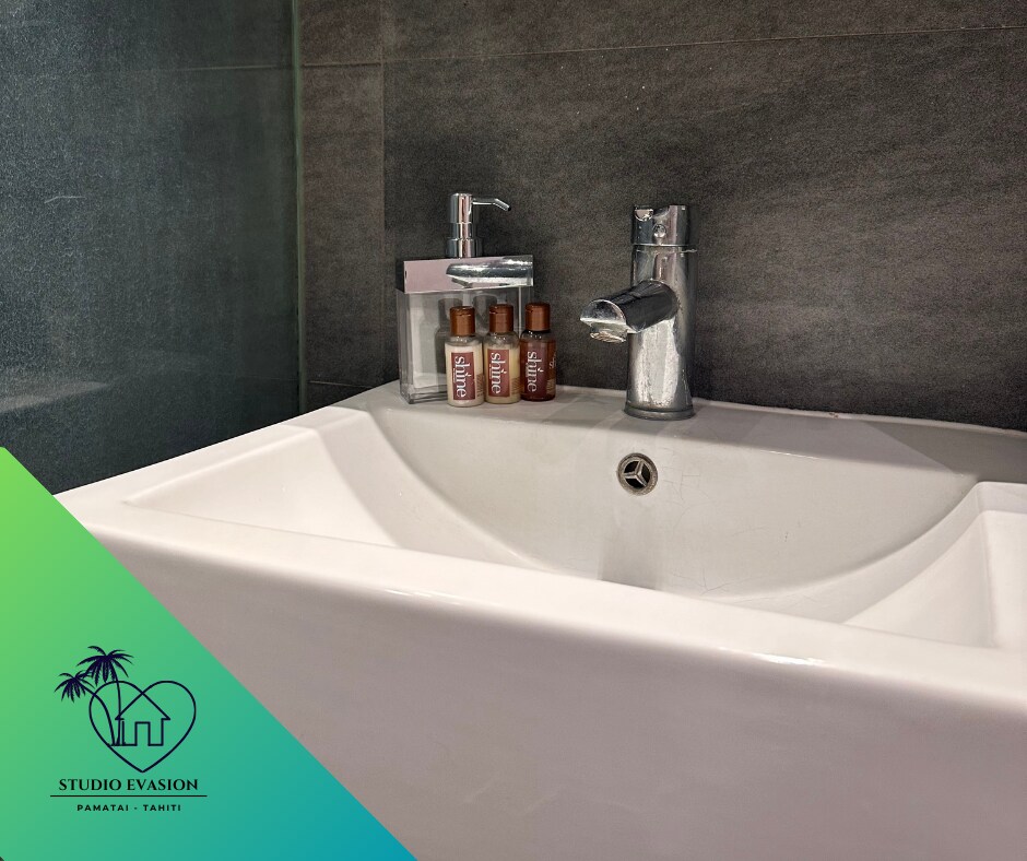 A modern sink is displayed against a sleek, dark background. Small bottles of toiletries are neatly arranged beside the faucet, which is polished and reflective. The overall design emphasizes cleanliness and functionality.