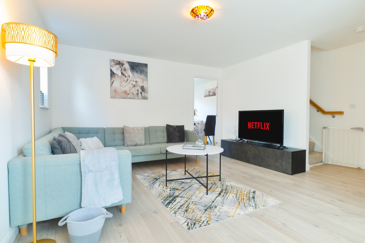 A bright living area is furnished with a light green couch and a coffee table atop a patterned rug. A flat-screen TV displays the Netflix logo, while natural light illuminates the space from nearby windows, complemented by a decorative lamp.