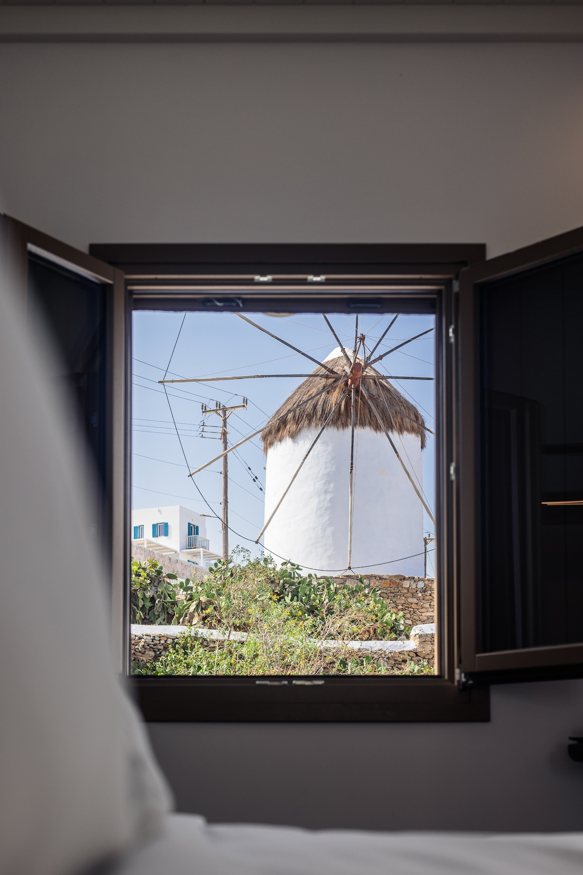 A window frame offers a clear view of the iconic Bonis Windmill, showcasing its distinctive thatched roof and towering structure. Surrounding greenery adds a touch of nature, while utility lines are visible in the background, providing an authentic glimpse of Mykonos town.