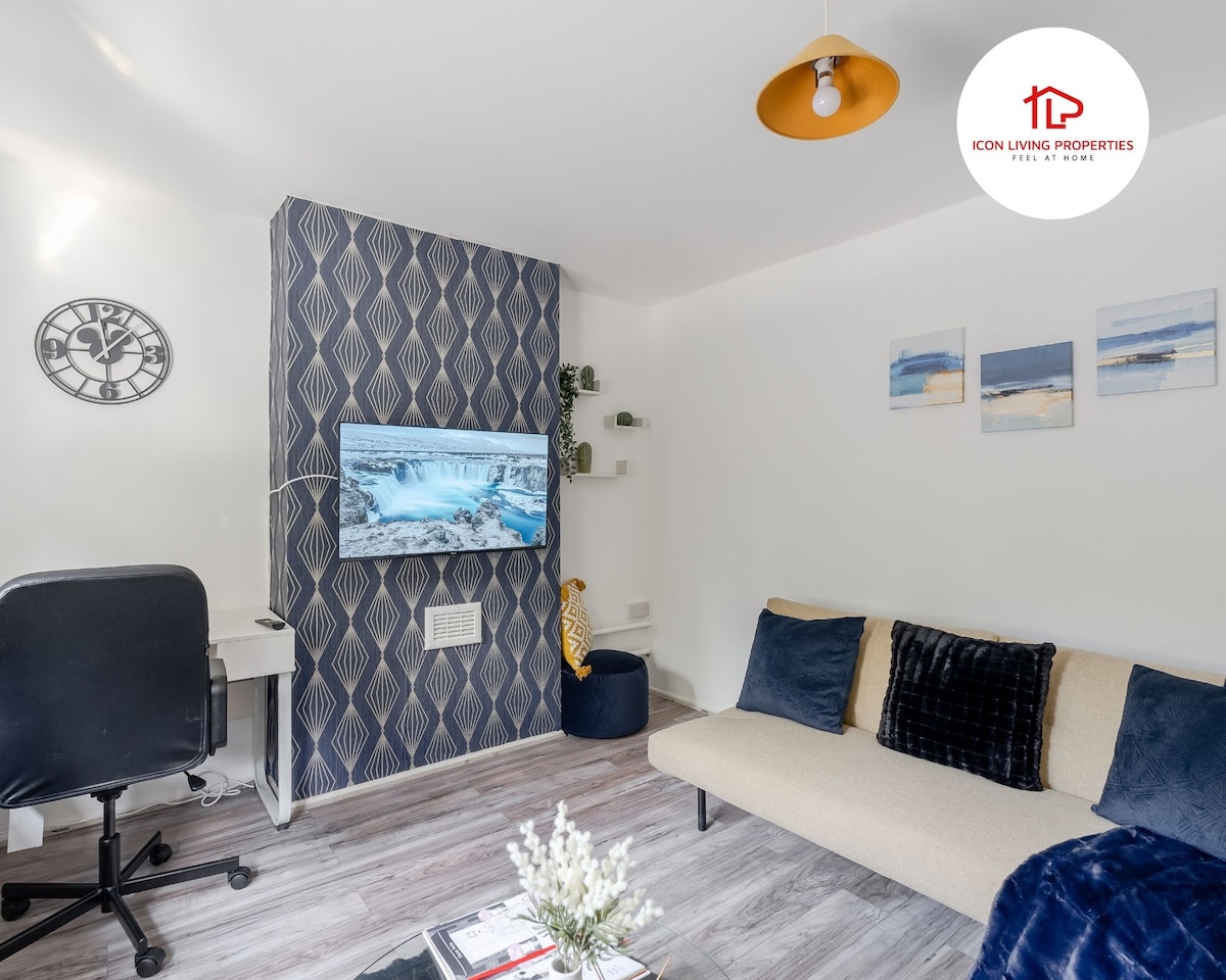 The living area features a modern sofa with plush cushions and a desk with a chair. A wall-mounted 40-inch smart TV displays a serene nature scene. A decorative clock and framed artwork are present, while the room is softly illuminated by a stylish pendant light.