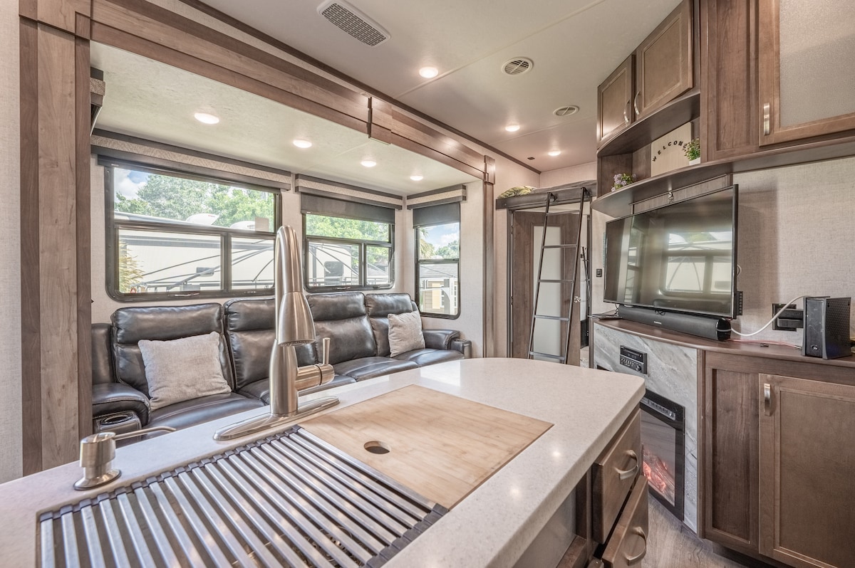 A well-equipped living area showcases a sleek kitchen island with a wooden cutting board and a recessed sink. Comfortable leather seating is arranged near large windows that provide ample natural light. A modern entertainment unit is visible, featuring a television and decorative plant.