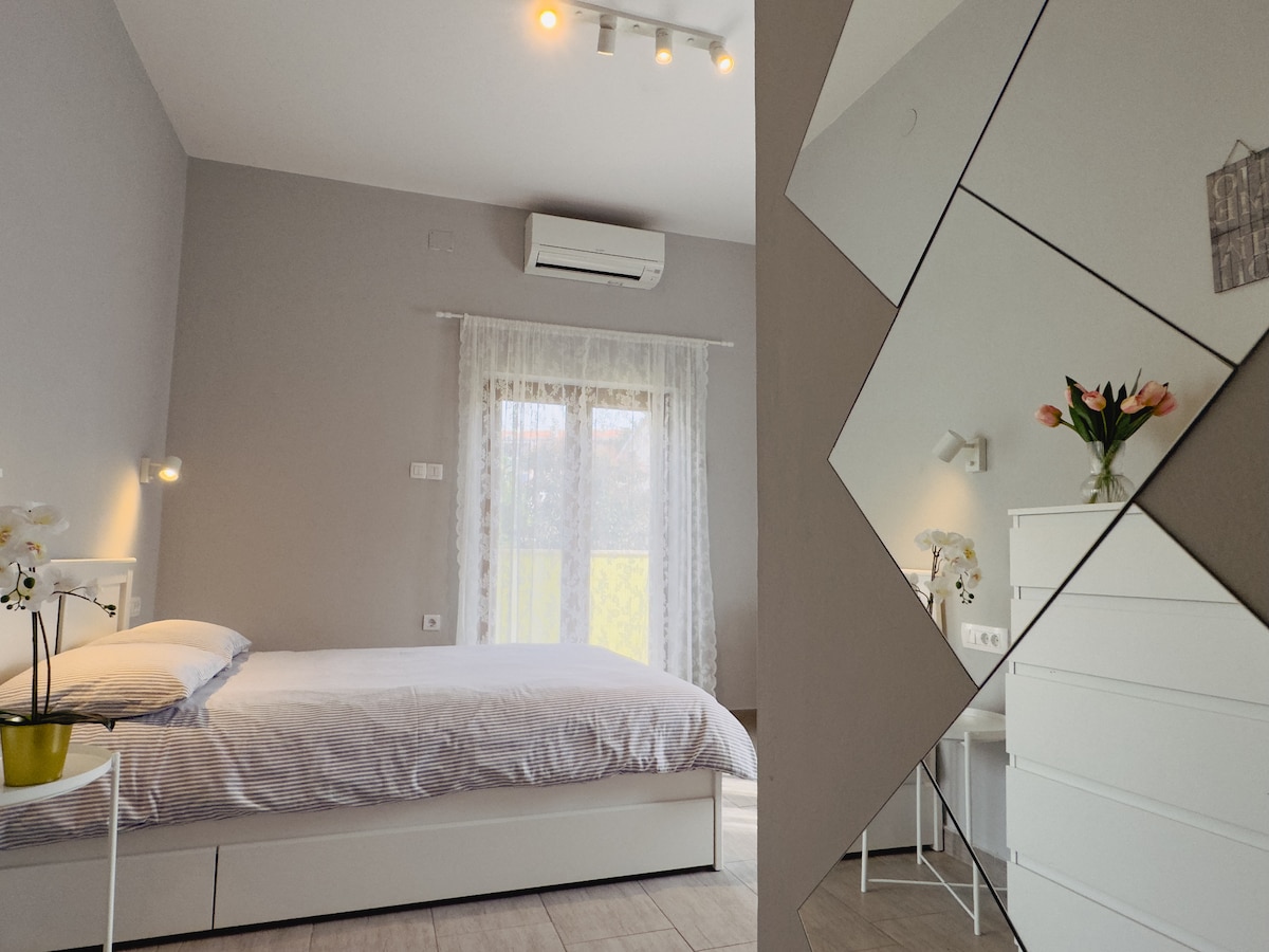 A bright sleeping area features a spacious bed with a striped duvet cover. Natural light enters through a curtain-draped window, illuminating the room. A decorative vase with flowers is seen on a dresser, while abstract mirrors reflect the modern design. Air conditioning is visible on the wall.