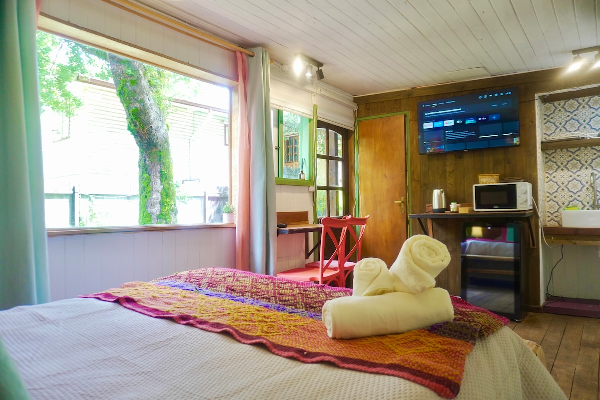 A cozy sleeping area features a spacious bed adorned with a colorful blanket. White towels are neatly arranged at the foot of the bed. Natural light fills the room through large windows, highlighting a small table and chair, as well as a kitchenette with a microwave and television.