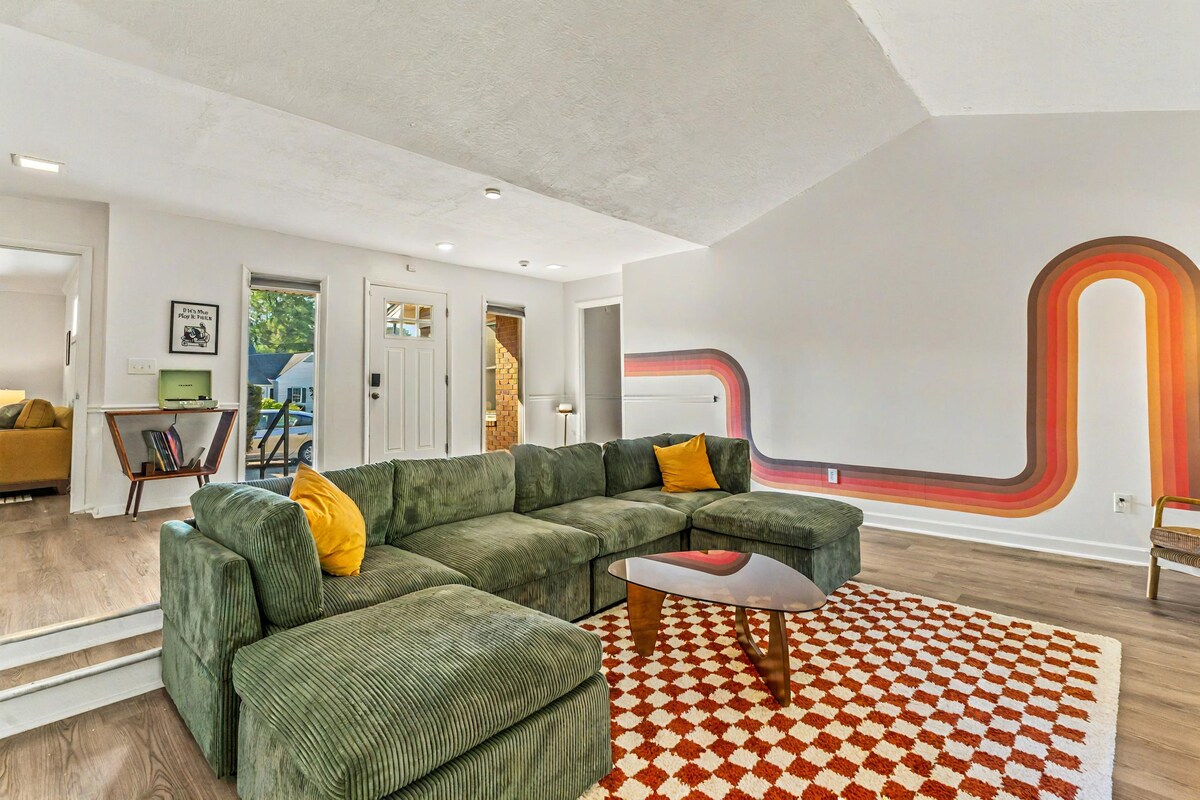 A spacious living area features a large green sectional sofa, accented with a mustard yellow pillow. A round coffee table sits on a patterned area rug. Natural light streams through the adjacent doors, highlighting the minimalist decor and unique wall art.
