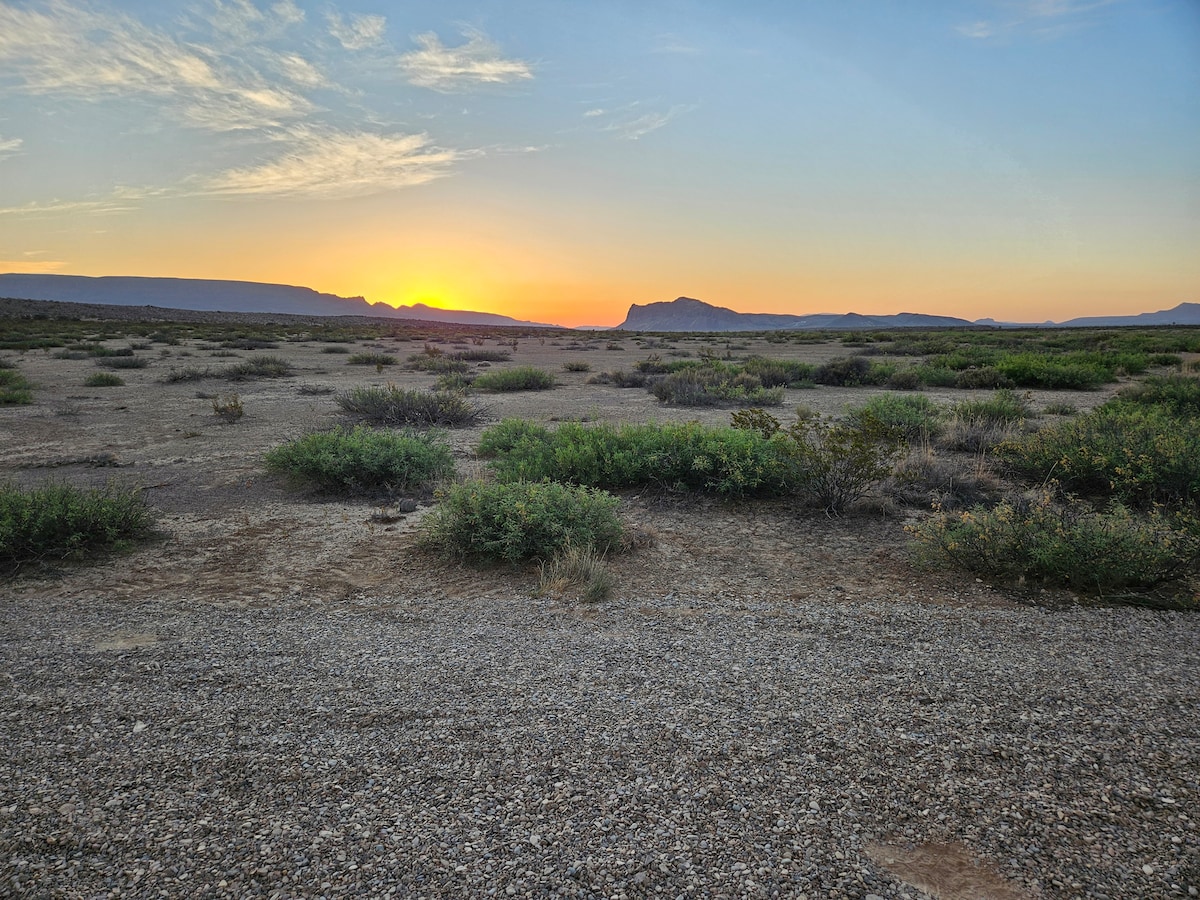 A serene desert landscape is illuminated by the warm hues of sunrise. Low vegetation and scattered shrubs are visible across the gravel expanse, while distant mountains form a backdrop against the colorful sky, signaling the start of a new day.