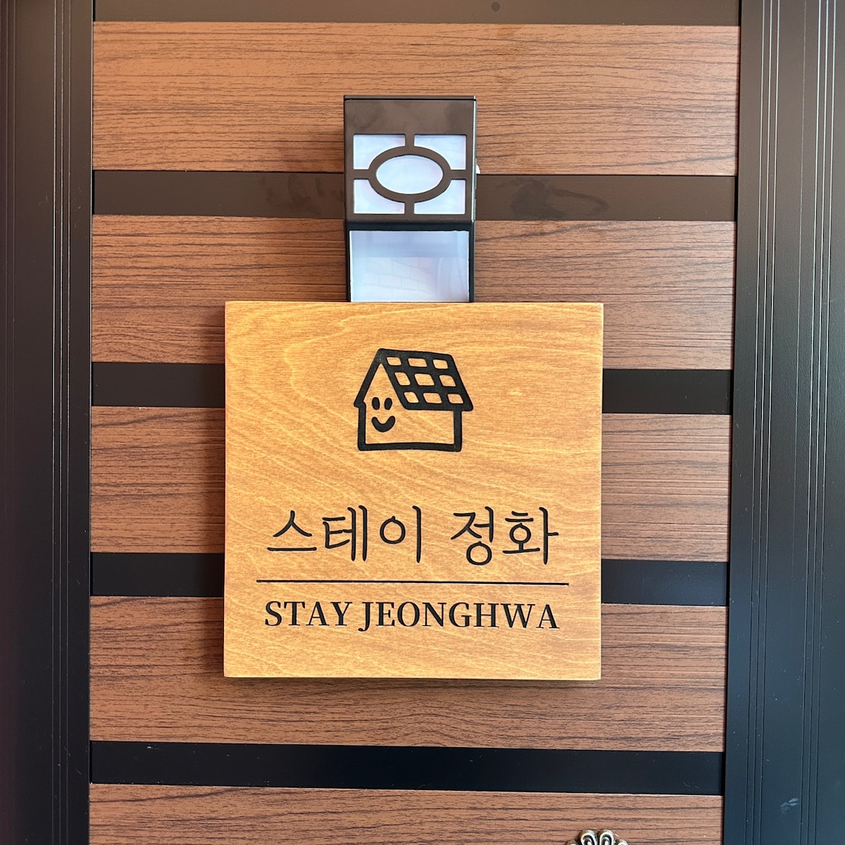 A wooden sign displaying the name 'STAY JEONGHWA' text in both English and Korean is mounted on a stylized wooden door. A simple house illustration and a welcoming smiley face are featured above the text, creating an inviting first impression.