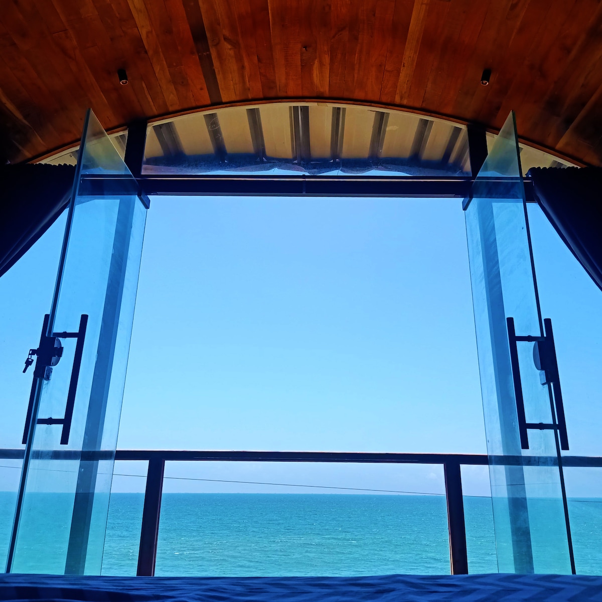 A panoramic view of the ocean is framed by two glass doors, partially opened to reveal the vibrant blue waters. The wooden ceiling adds warmth to the space, while the smooth horizon meets a clear sky.