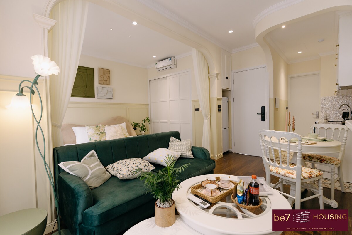 A living area is depicted with a green sofa adorned with decorative pillows. A round coffee table occupies the center, while a small dining table is positioned to the right. Natural light illuminates the space, highlighting the soft color palette and textured elements of the decor.