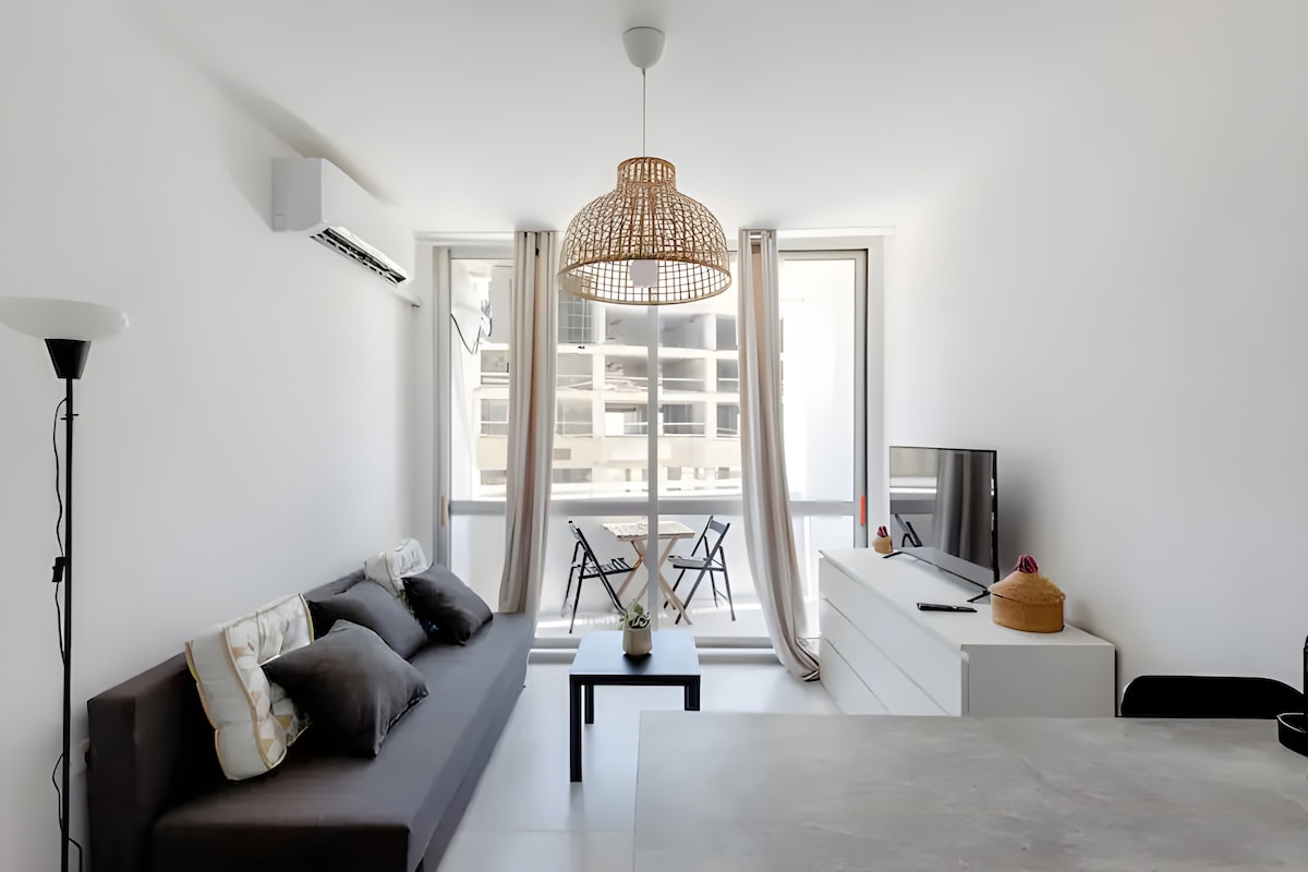 A modern living area is presented with a light-colored sofa, accented by two dark cushions. A large window offers natural light, with a view of two balcony chairs. A flat-screen television sits atop a sleek white cabinet, enhancing the clean aesthetic.